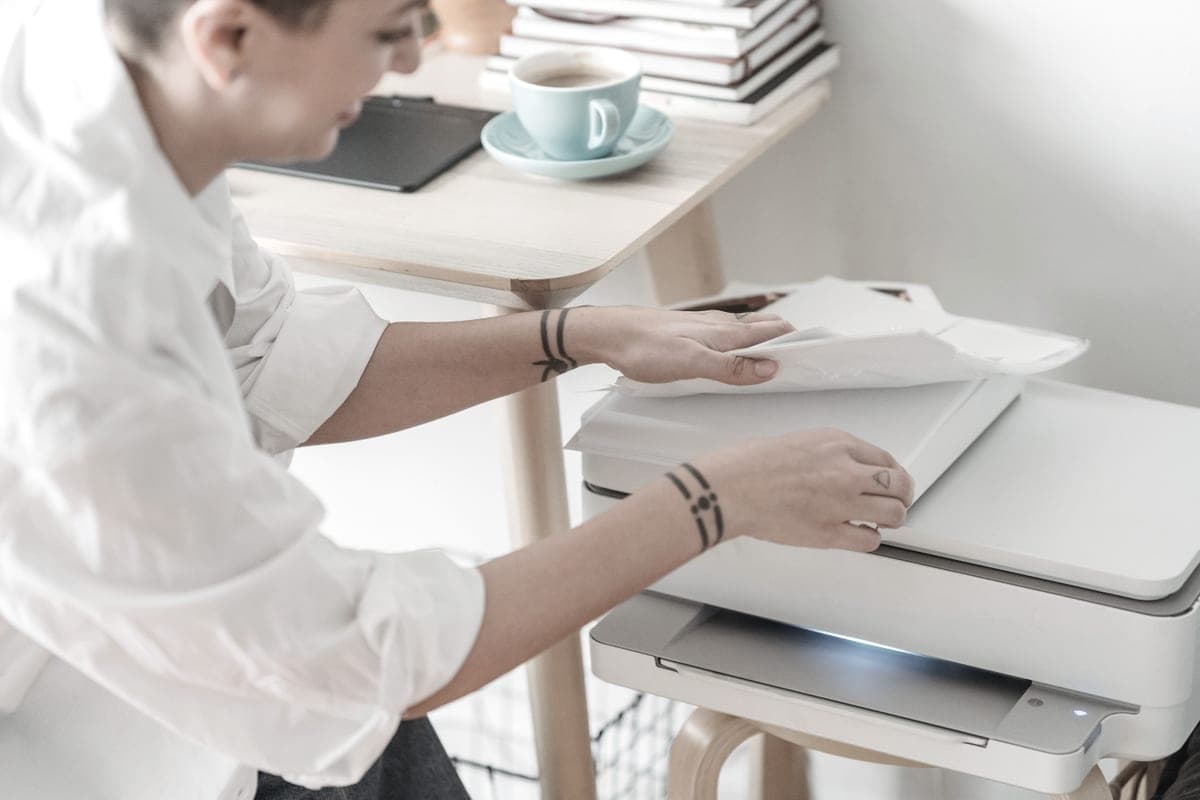 A hand lifting sheets of plain paper beside a printer while working from home